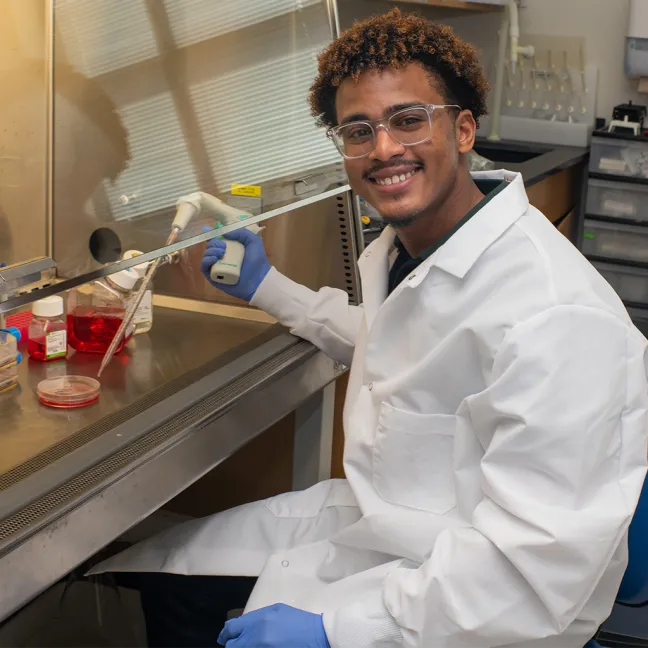 A U N E student in a lab coat smiles while using a pipette at a laboratory workstation.