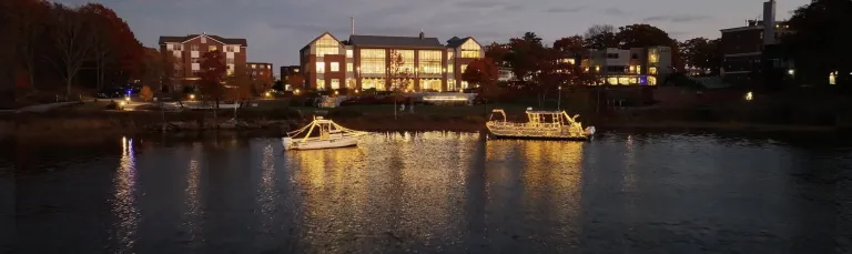 Two boats covered in white holiday lights sit in the water in front of the Ripich Commons on U N E's Biddeford campus
