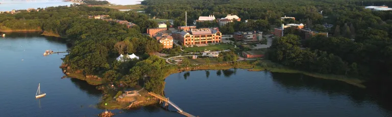 Aerial view of University of New England's Biddeford campus overlooking a calm cove. Red brick buildings sit on landscaped grounds surrounded by forest, with a small boat in the water and historic stone ruins visible along the shoreline.