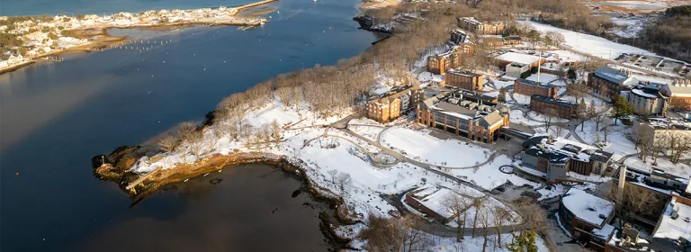 Aerial winter view of U N E's waterfront campus showing snow-covered grounds, brick academic buildings, residence halls, and harbor with coastal community in background.