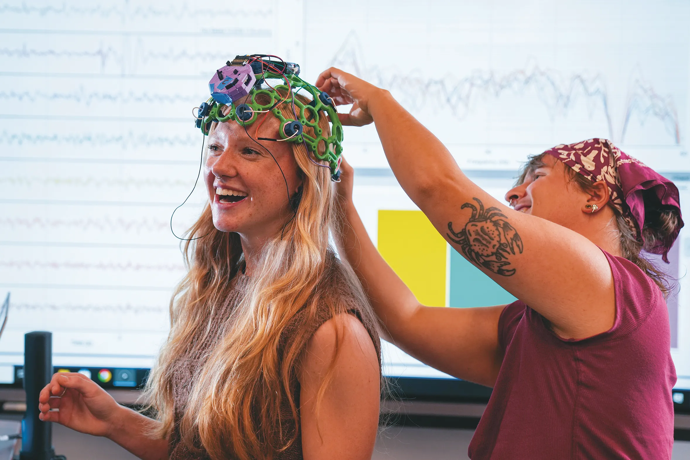 Milo Lypps adjusts a colorful wired skull caps on Jessica Howard's head in a laboratory or classroom setting.