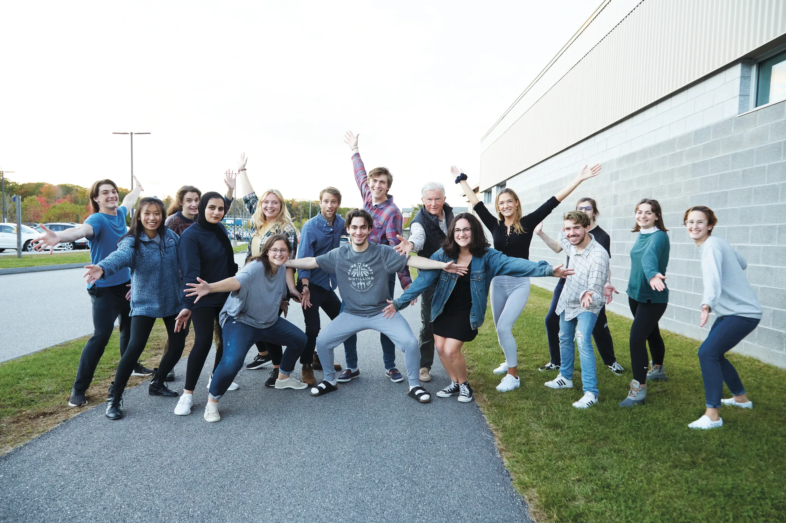 A diverse group of approximately 15 Shaw Innovation Fellows pose enthusiastically outside a modern UNE building, with students striking playful poses with arms raised and outstretched against an autumn campus backdrop