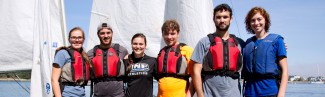 A group of students pose wearing life vests in front of a sailboat
