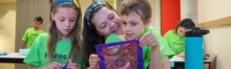 A U N E student compliments two children's artworks made during Autism Camp
