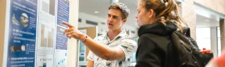 A student points to their presentation poster as they talk to a fellow student