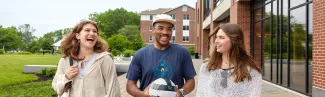 Three U N E students laugh together as they walk through Biddeford Campus, with a brick building to their right and green grass on their left