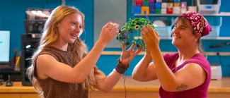 Two students examine a colorful 3D-printed EEG sensor cap standing at wooden table in innovation lab with blue shelving and equipment behind them.