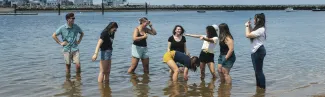A group of students laugh and play in the shallow water along a sunny coastal shoreline, with a rocky seawall, marina, and New England waterfront homes visible in the background.