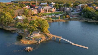 Aerial view of UNE's Biddeford campus in autumn, showing the waterfront campus with brick academic buildings, a small island with pavilion connected by a wooden footbridge, surrounded by colorful fall foliage and the Saco River estuary.