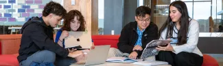 Four students study together on red curved seating around a round table with laptops and textbooks in a modern campus building with colorful wall displays.