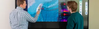 A U N E instructor points to seismic and geographic data on a large touchscreen display while a student looks on.
