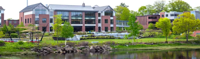 A large brick building with large windows sits on the edge of a river bank surrounded by green trees and grass