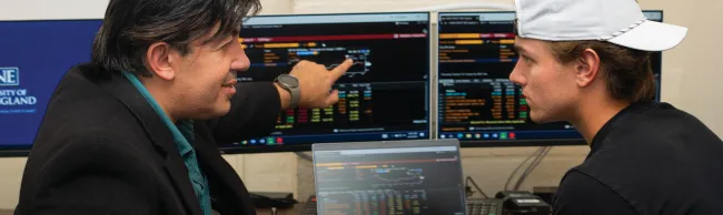 A business professor talks about finance to a student while pointing to dual monitors that display financial data