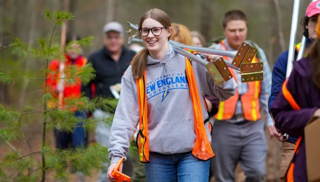 A UNE student carries research equipment through the woods