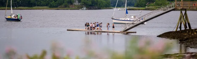 A group of students walks along a floating dock on the Saco River near UNE's Biddeford campus, with sailboats and a wooded shoreline in the background.