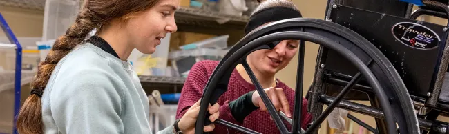 Two U N E students work together to repair a wheelchair wheel in an assistive technology lab.