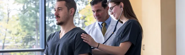A U N E Physician Assistant student listens to a classmate's back with a stethoscope while an instructor observes.