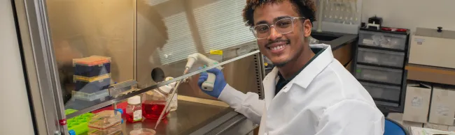 A U N E student in a lab coat smiles while using a pipette at a laboratory workstation.