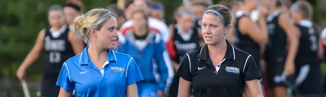 Two U N E field hockey coaches walk together with the team in the background before a game.