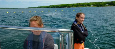 A female student stands at the back of a UNE boat while another female student pilots the boat