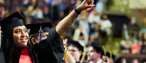 A woman in a cap and gown points to friends at UNE's 187th Commencement ceremony
