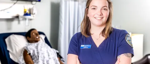 Portrait of Anne Carrigg, a nursing student, in UNE's simulation lab. A patient simulator is behind her.