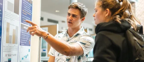 A student points to their presentation poster as they talk to a fellow student