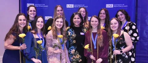 A group of students and faculty pose for a photo against a UNE-branded step-and-repeat