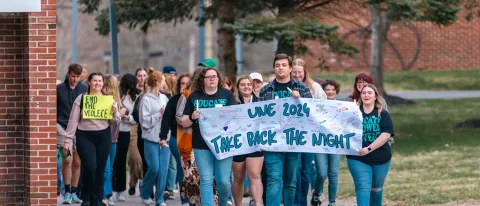 A group of students marches on campus holding a sign that reads "UNE 2024 Take Back the Night"