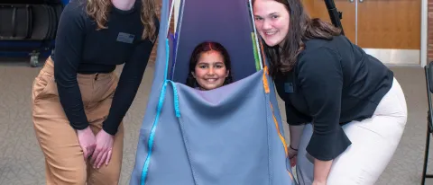 A child sits inside a suspended tent and two students pose around her