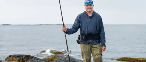 A UNE student poses with a fishing rod in front of the ocean