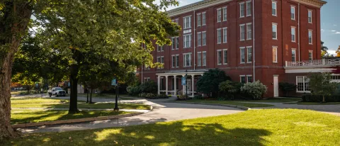 A brick building sits behind green grass and a large tree