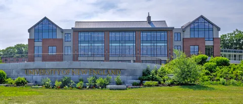 A brick building with numerous windows sits behind a stone sign that reads, "University of New England"