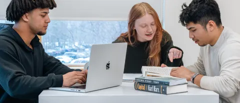 A student types on their laptop while two students review a book while sitting together at a high-top desk in front of a large window