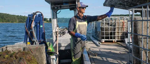 A student wearing waiters and a U N E baseball cap props up an item on a boat