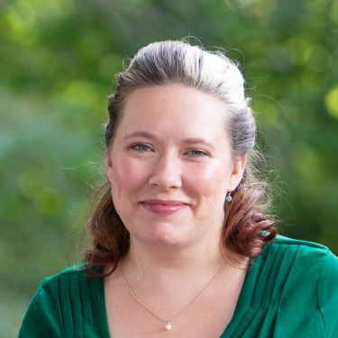 Cera is smiling at the camera with soft makeup, her hair is in an updo, and she is wearing a green dress.