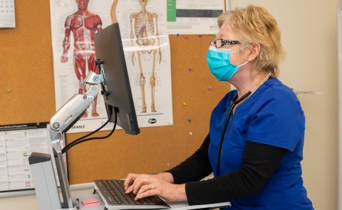Woman with blonde hair in scrubs stands at computer in campus health center