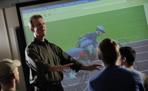 UNE Professor Jim Cavanaugh teaches students in front of a screen with cyclists in view