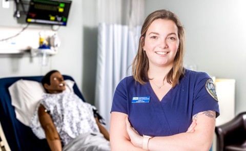 Portrait of Anne Carrigg, a nursing student, in UNE's simulation lab. A patient simulator is behind her.