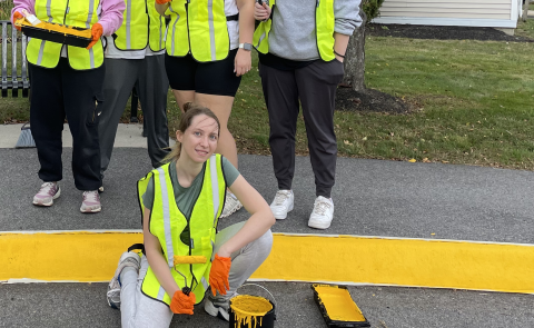 A group of students wearing yellow safety vests pose in front of the Westbrook Housing building displaying the painted sidewalk curbs they completed to enhance safety