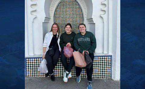 Three female students pose in front of Moroccan architecture. Photo is set against a blue background.