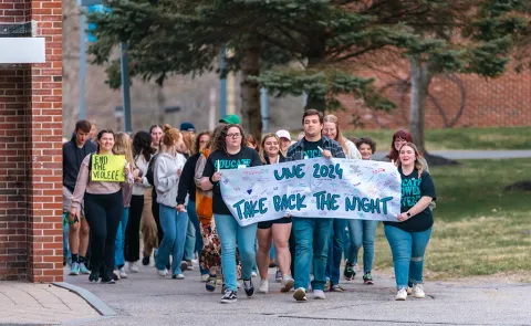 A group of students marches on campus holding a sign that reads "UNE 2024 Take Back the Night"