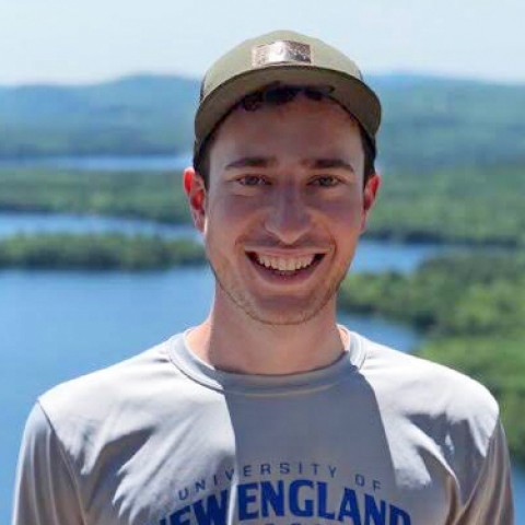 Mitchell Becker stands smiling at the camera atop a mountain. We see blue lakes and green mountains in the background