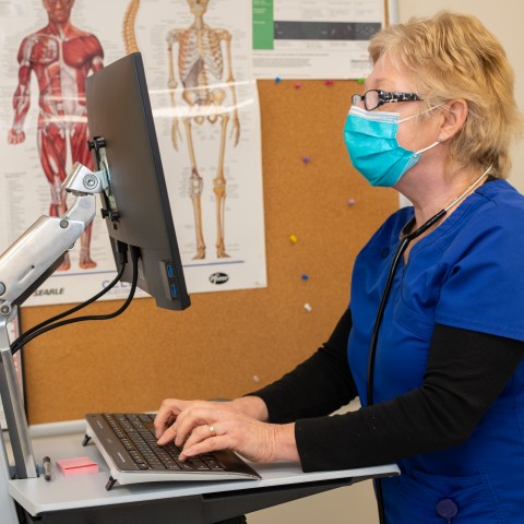 Woman with blonde hair in scrubs stands at computer in campus health center
