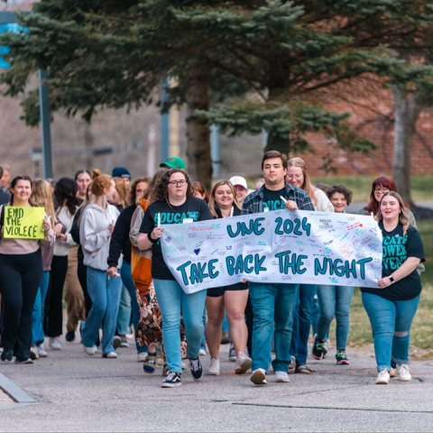 A group of students marches on campus holding a sign that reads "UNE 2024 Take Back the Night"