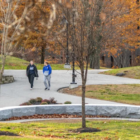 Two U N E students walk on a paved path through the greenery of Biddeford Campus