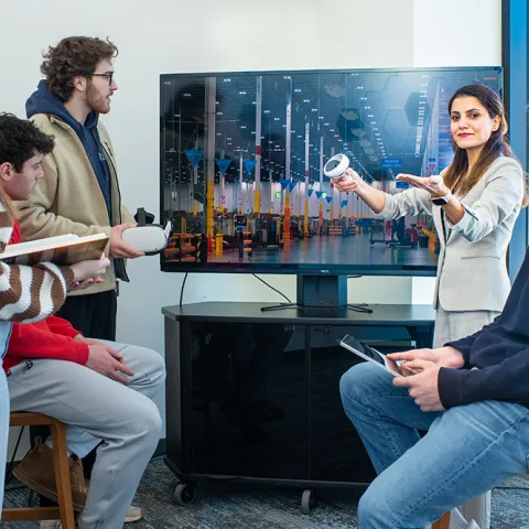 A group of four students listen intently to their professor as she describes a virtual warehouse image on a large monitor