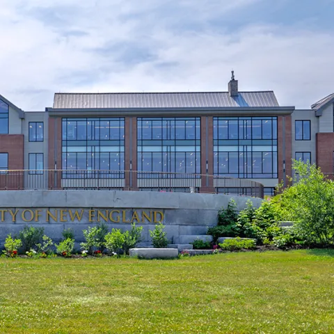 A brick building with numerous windows sits behind a stone sign that reads, "University of New England"