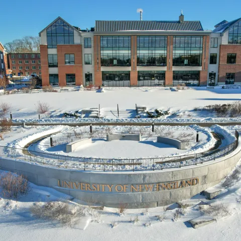 Circular stone amphitheater with 'University of New England' inscription in foreground, modern glass-fronted academic building and residence halls visible across snowy landscape.