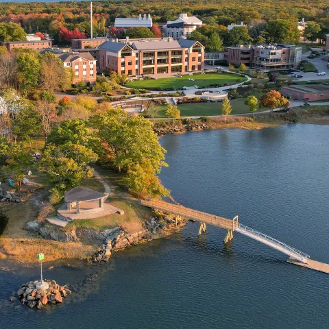 Aerial view of UNE's Biddeford campus in autumn, showing the waterfront campus with brick academic buildings, a small island with pavilion connected by a wooden footbridge, surrounded by colorful fall foliage and the Saco River estuary.